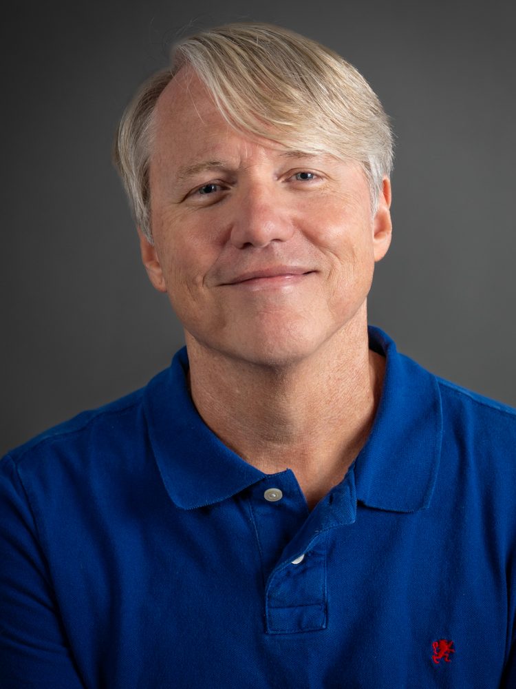 A headshot of Kenneth Kidd. He is wearing a blue and orange polo shirt and smiling. His light hair is parted to the side.