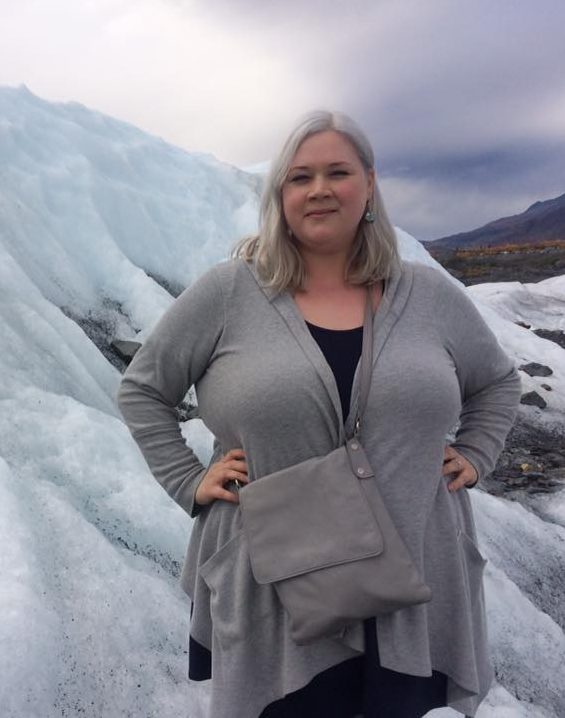 K. Blasco Soler smiles with hands on hips wearing black and gray with a bag slung over their shoulder. In the near background: Matanuska Glacier, in the far background: the Chugach Mountains in Alaska.