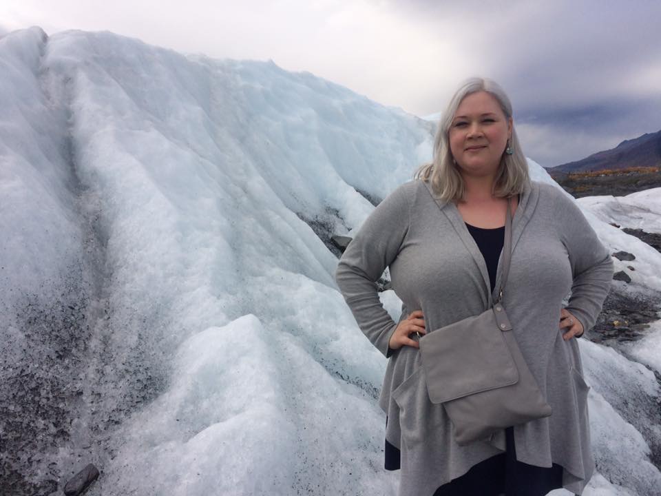 K Blasco Soler stands beside a pale blue and silty glacial formation atop Atop Matanuska Glacier. K is smiling with hands on hips and wearing a gray sweater and shoulder bag.