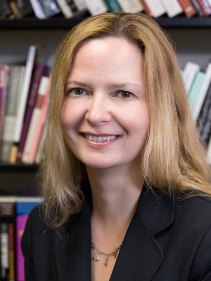Ange Mlinko smiling, seated in front of bookshelves. She has long, light colored hair and is wearing a black blazer.