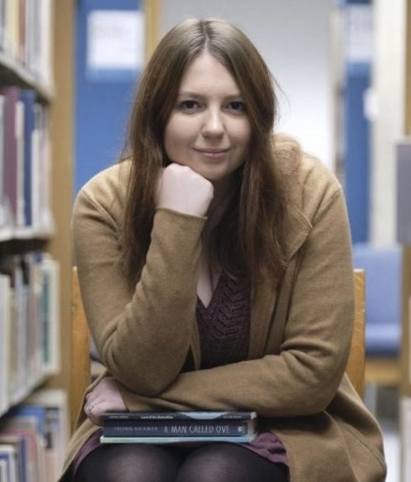 Chloe Cook sits in the middle of a library aisle. She is facing the camera straight-on, with her jaw resting on her fist
