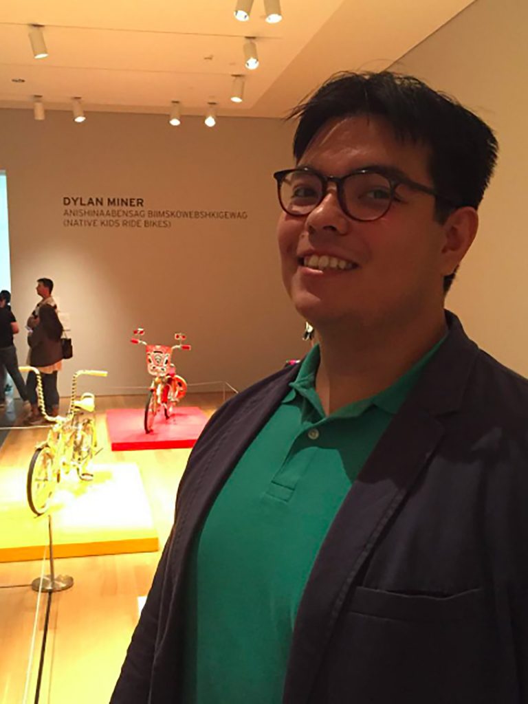 Victor Del Hierro smiling in front of an indoor art exhibit by Dylan Miner. He is wearing a green shirt, black blazer, and black glasses.