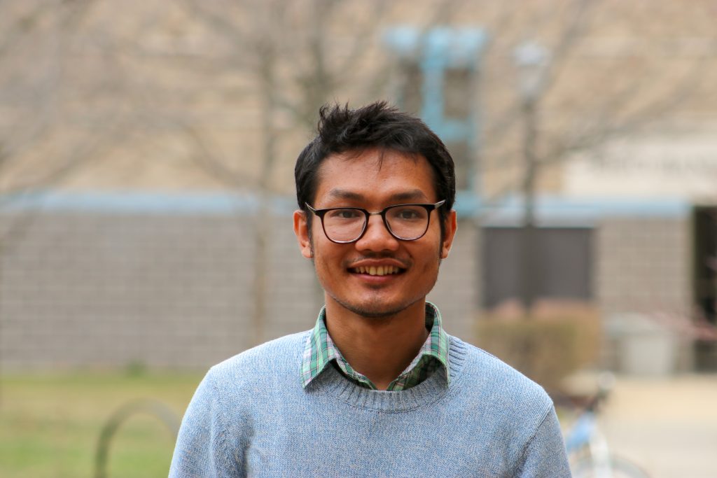 Dinalo Chakma smiles at the camera. He wears a light blue sweater over a plaid green and white collared shirt, as well as black framed glasses.