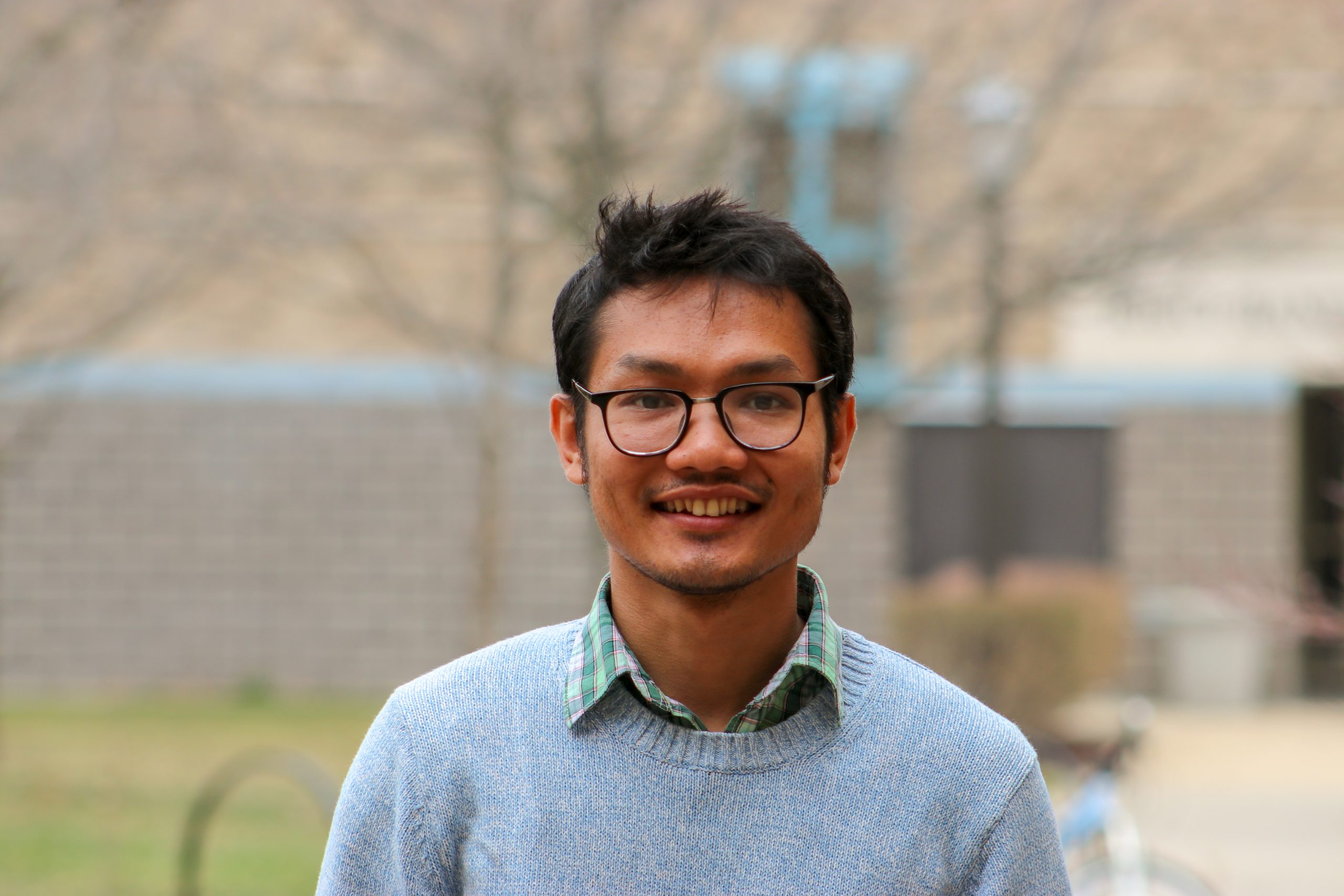 Dinalo Chakma smiles at the camera. He wears a light blue sweater over a plaid green and white collared shirt, as well as black framed glasses.