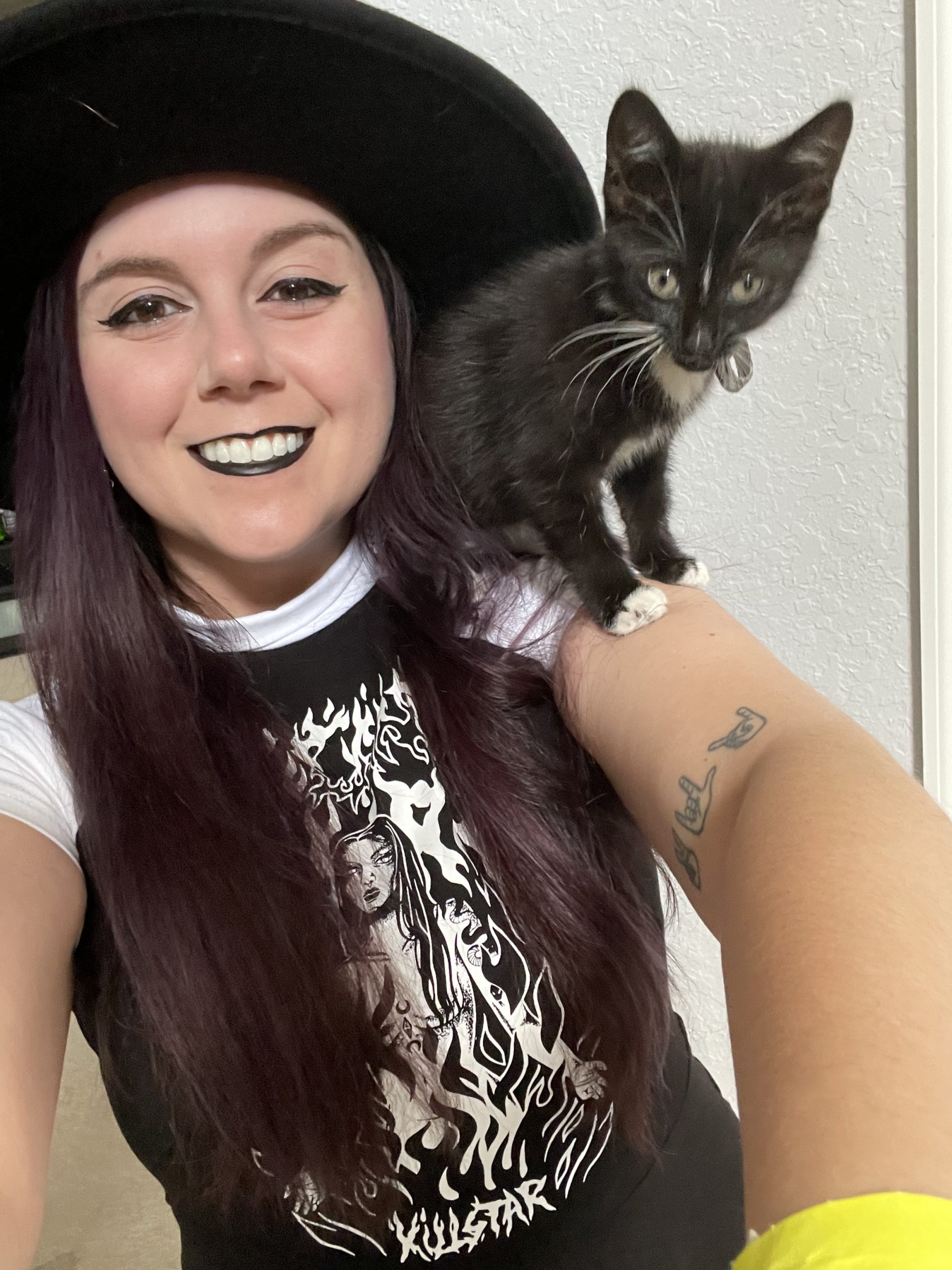 A black-haired woman with a tiny tuxedo kitten on her shoulder smiles at the camera.