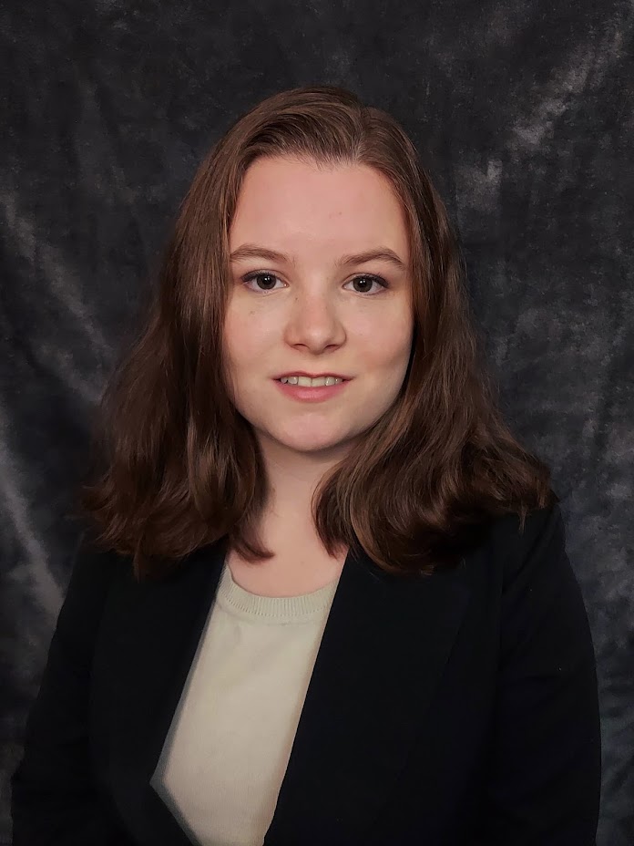  Emily faces the camera. She wears a black blazer and stands in front of a gray background.