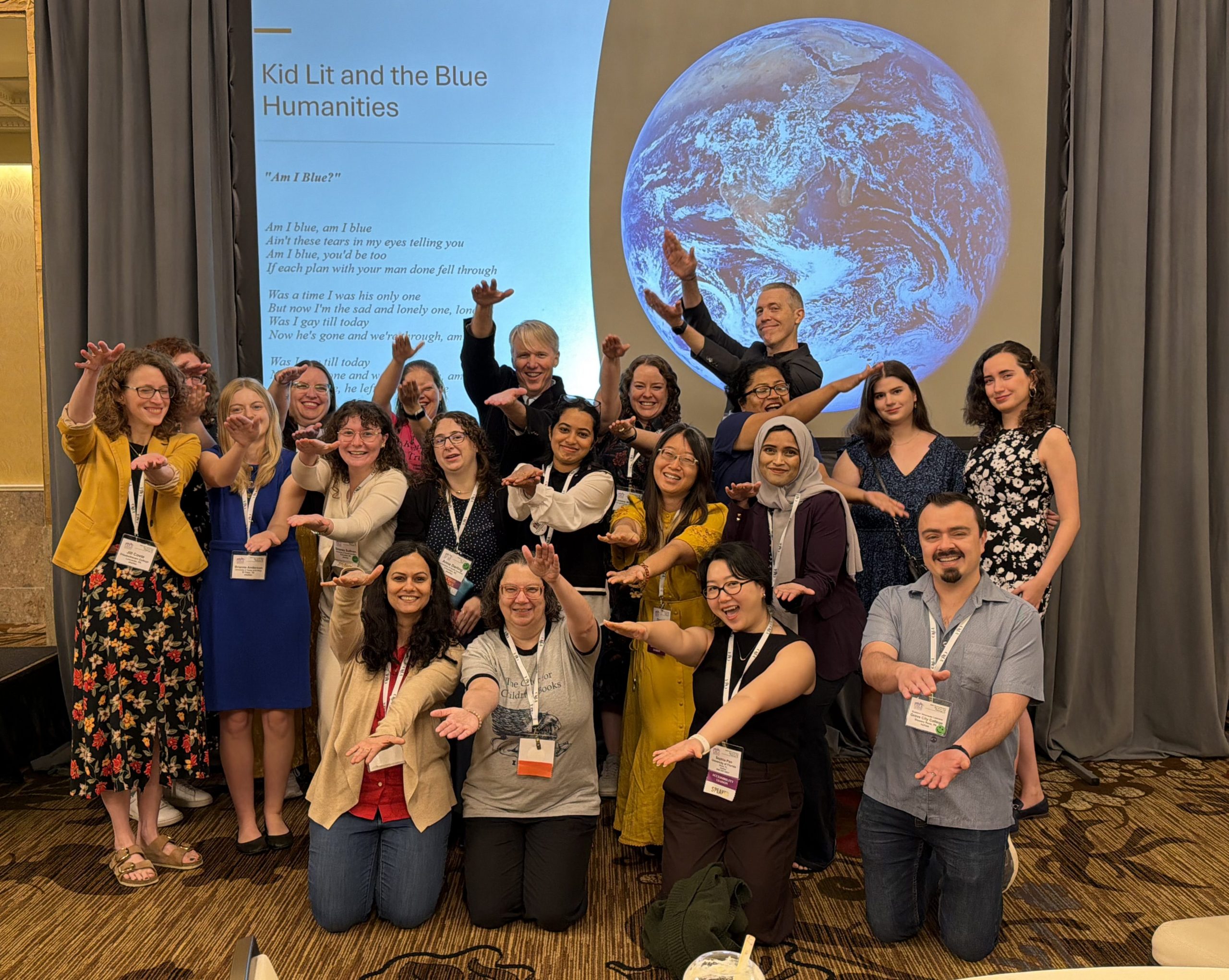 Around 20 people, including Kenneth Kidd, graduate students, and alumni, gathered in front of a conference presentation slide. They are doing the Gator chomp with arms outstretched.