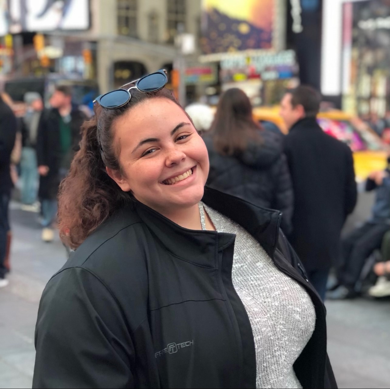 Judy smiles at the camera. She is wearing a grey sweater and black jacket. The background is a blurred city street.