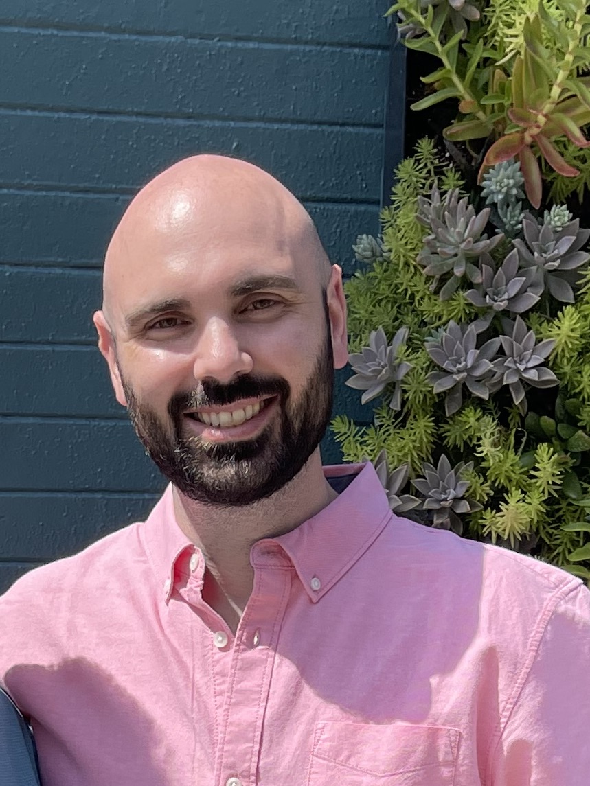Kevin smiles at the camera in a pink polo shirt in front of a navy blue wall.