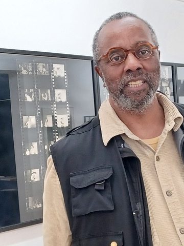 Dr. Mark Reid stands in front of a wall of framed cinema film negatives. He is wearing a tan dress shirt, black vest, and brown-rimmed glasses, and he is smiling at the camera.