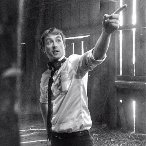 A photo of Matt on the set of his first feature film as writer/director. The image is in black and white. Matt wears a white button down shirt with the sleeves rolled up to his elbows and a black tie. He is pointing off camera.