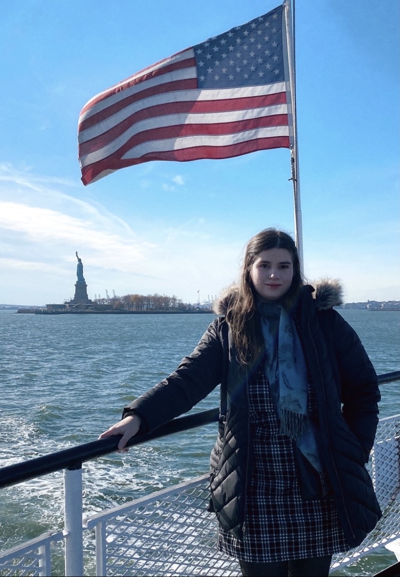 Natalie is wearing winter clothing, standing on a boat with the statue of liberty and an American flag in the background.