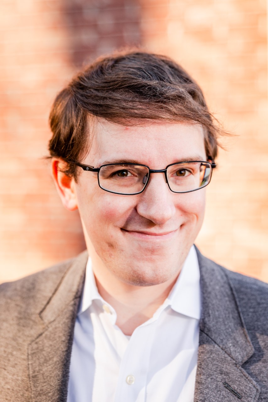 Ryan Kerr smiles at the camera. He is wearing a white dress-shirt, gray blazer, and glasses with black frames.