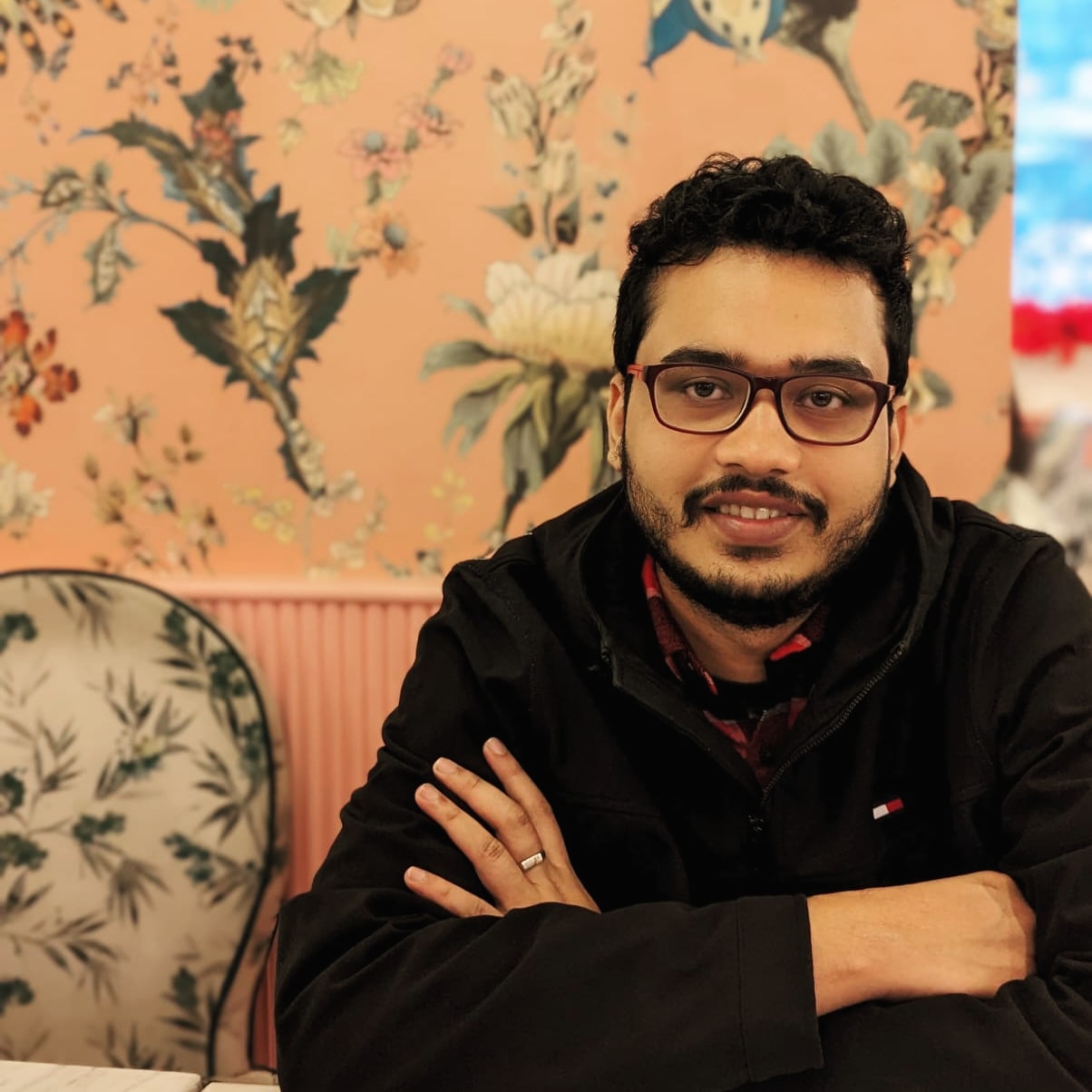 Suvendu Ghatak sits against a floral printed background, wearing a black jacket, and smiles at the camera.