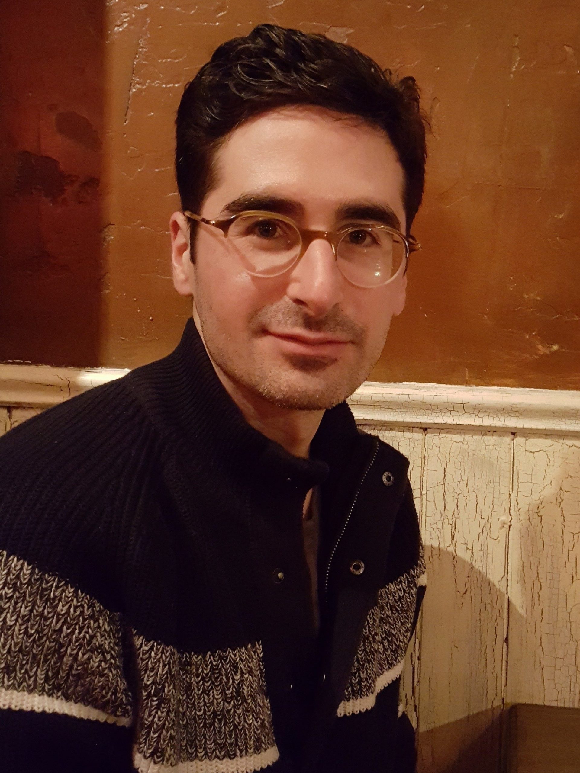 A photo of Trevor Mowchun wearing a dark, striped cardigan and glasses.