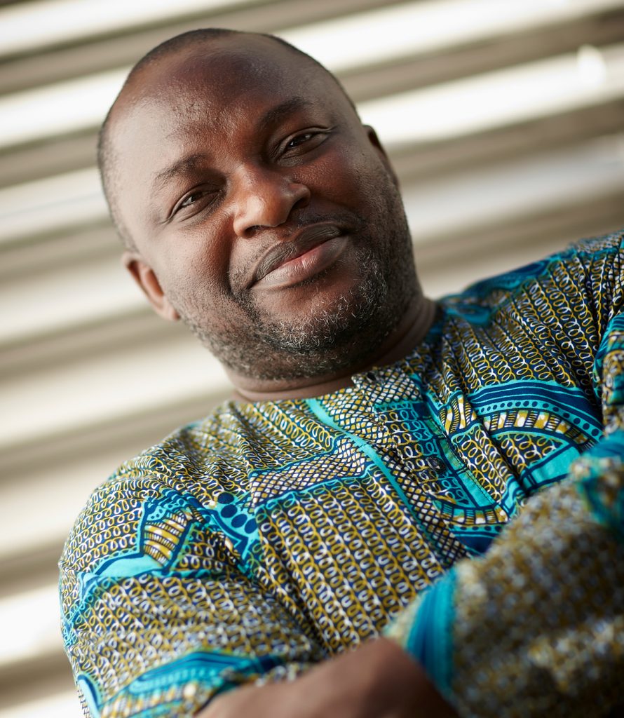 Uwem Akpan smiling with his arms crossed. He is wearing a colorful tunic.