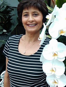 A photograph of Malini Schueller smiling broadly. She is holding a white orchid and surrounded by foliage.