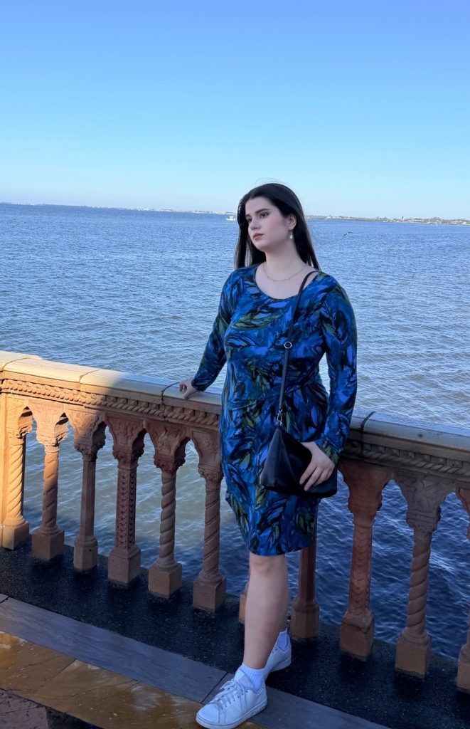 Natalie is standing on a waterfront wearing a blue dress. She is looking away from the camera.