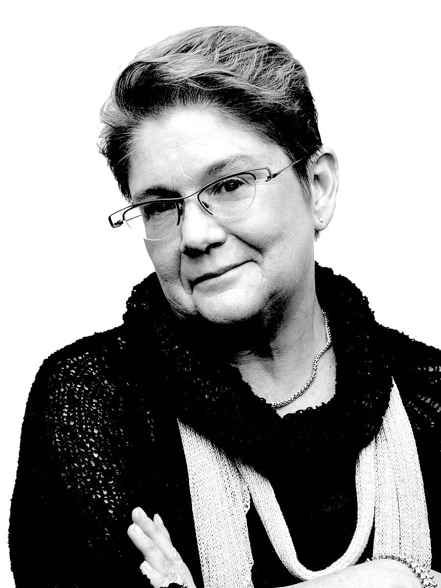 A black and white headshot of Stephanie Smith. Her head tilted to the side, she is smiling with her arms crossed. She is wearing layered scarves and glasses.