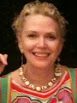 A photograph of Tace Hedrick smiling broadly. She is wearing a colorful blouse and light jewelry.
