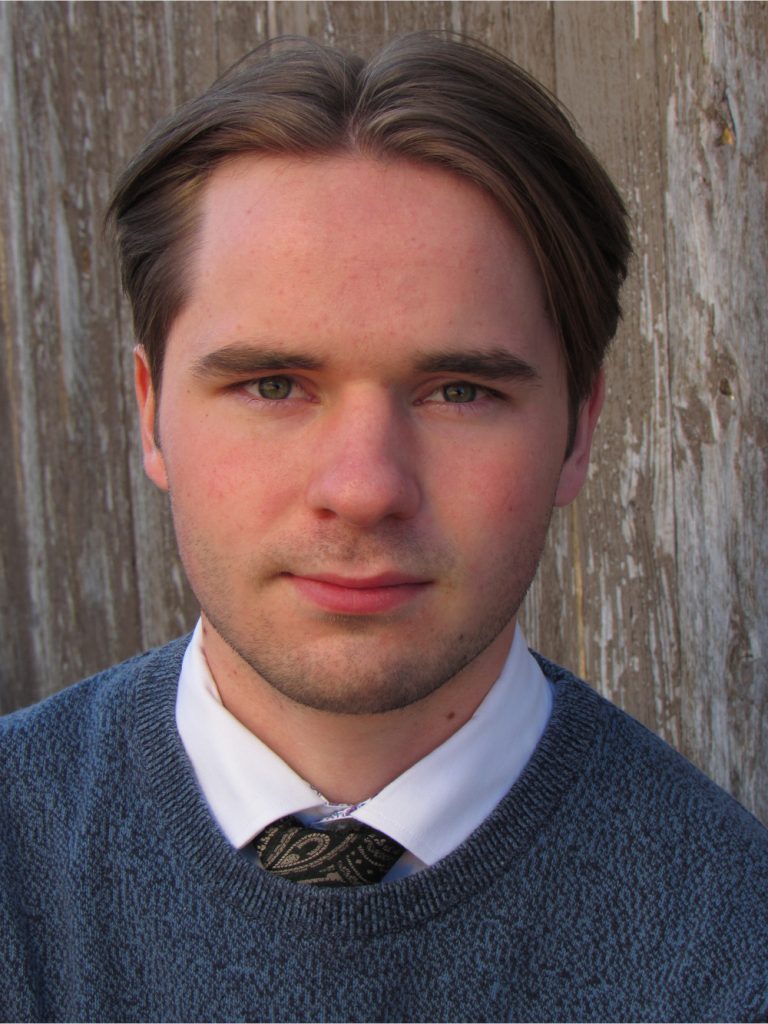 Thomas smiles at the camera. He is wearing a blue sweater over a white collared shirt and a green paisley tie.