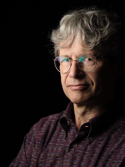 A headshot of William Logan. The background of the image is black, with his face and torso partially in shadow. He is wearing a button-down shirt and glasses.