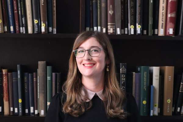 Adrienne Johnson is standing in front of a bookshelf in the Price Library of Judaica Suite. They are smiling warmly, with long hair parted to the side. They wear glasses and a black and white top with a peter pan collar.