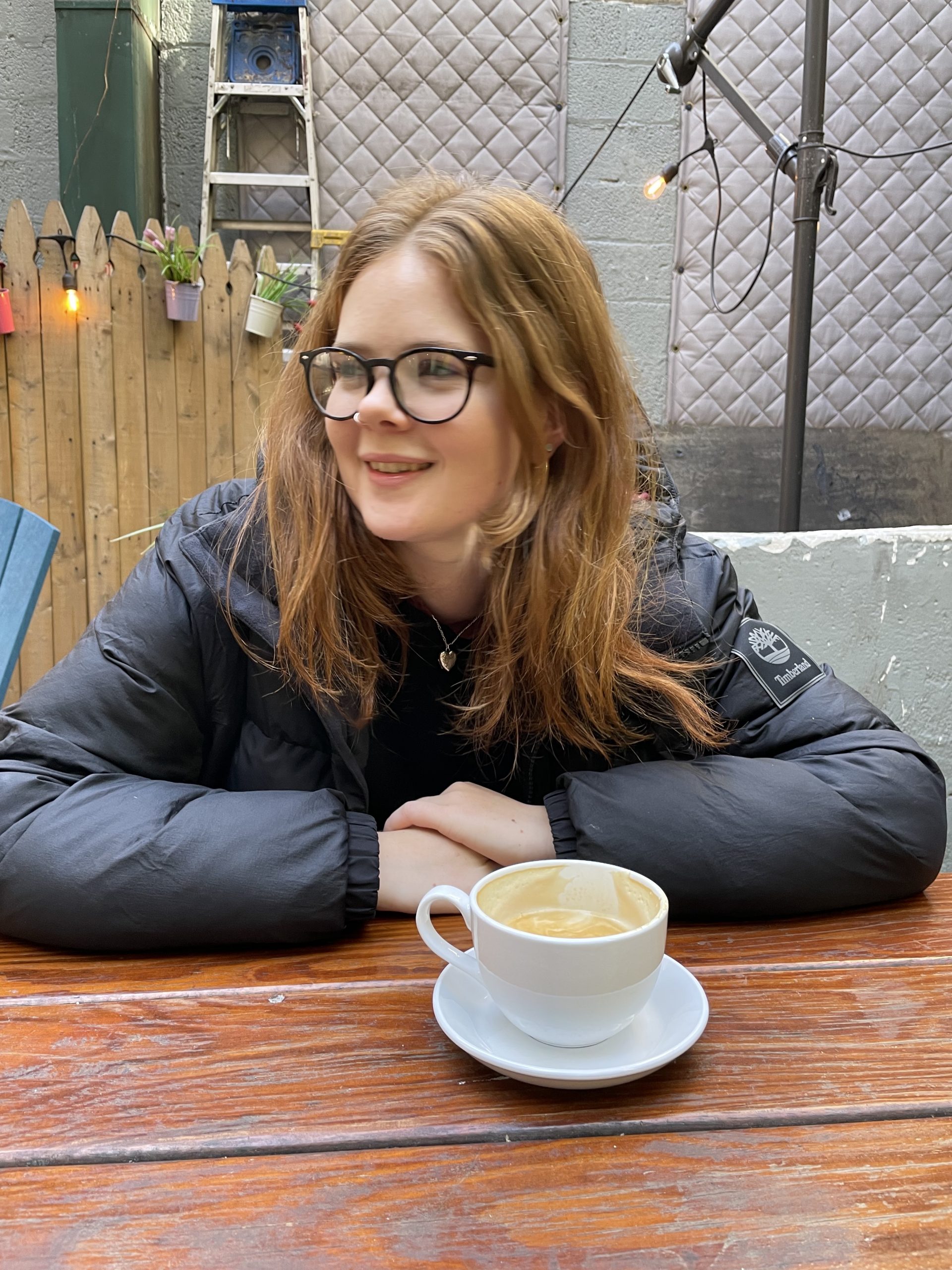 Albertine Clarke, a woman with red hair, sits at a wooden table with a cappuccino. She is wearing a black puffer jacket and glasses.