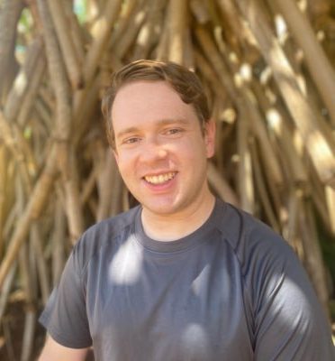 Alexander smiles at the camera. He is a white man wearing a grey T-shirt and standing in front of large tree roots.