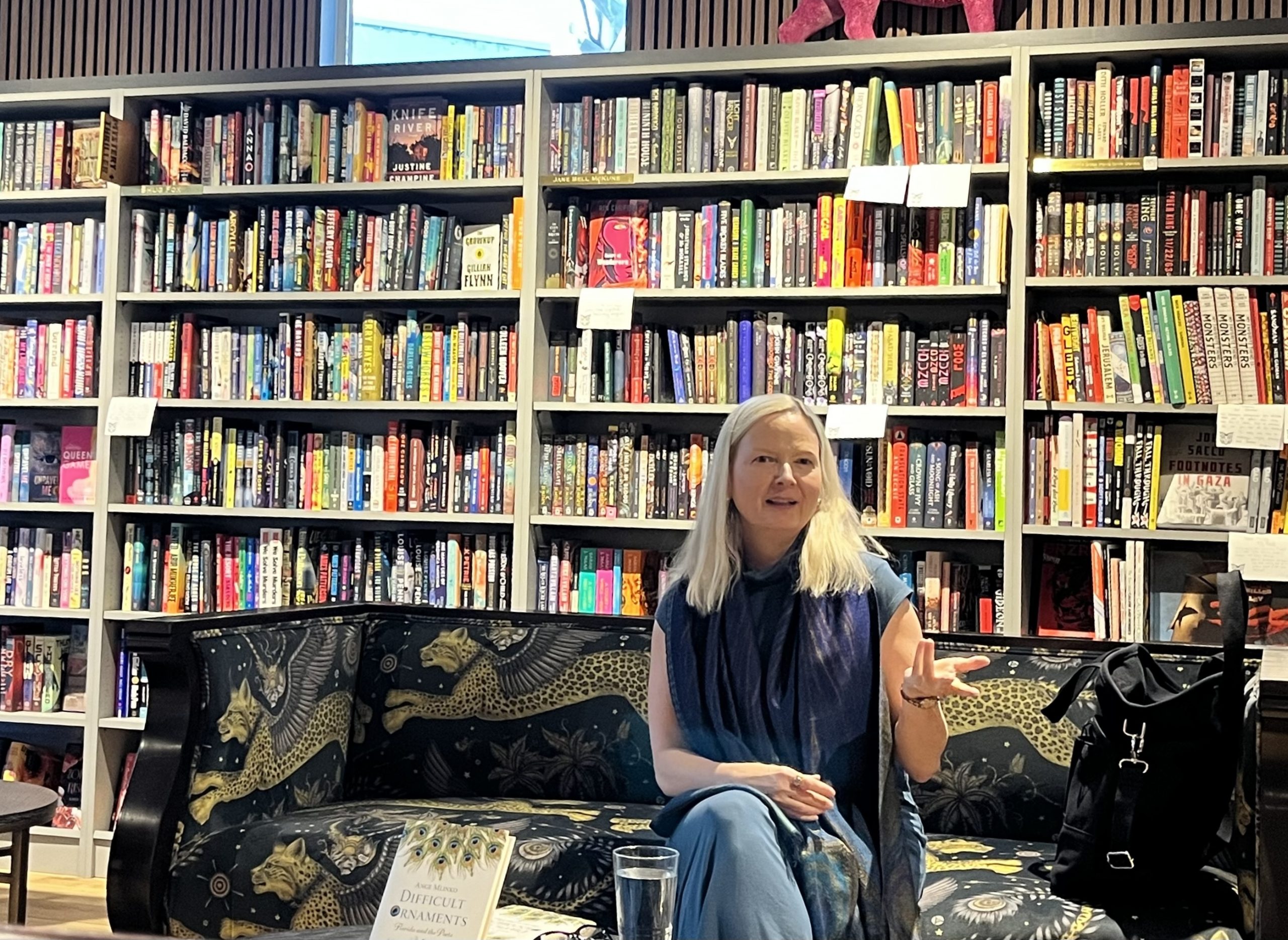 Ange Mlinko sits on a couch decorated in winged leopards. Behind her is walls of bookshelves. She is gesturing with her hand as though pictured mid-speech.