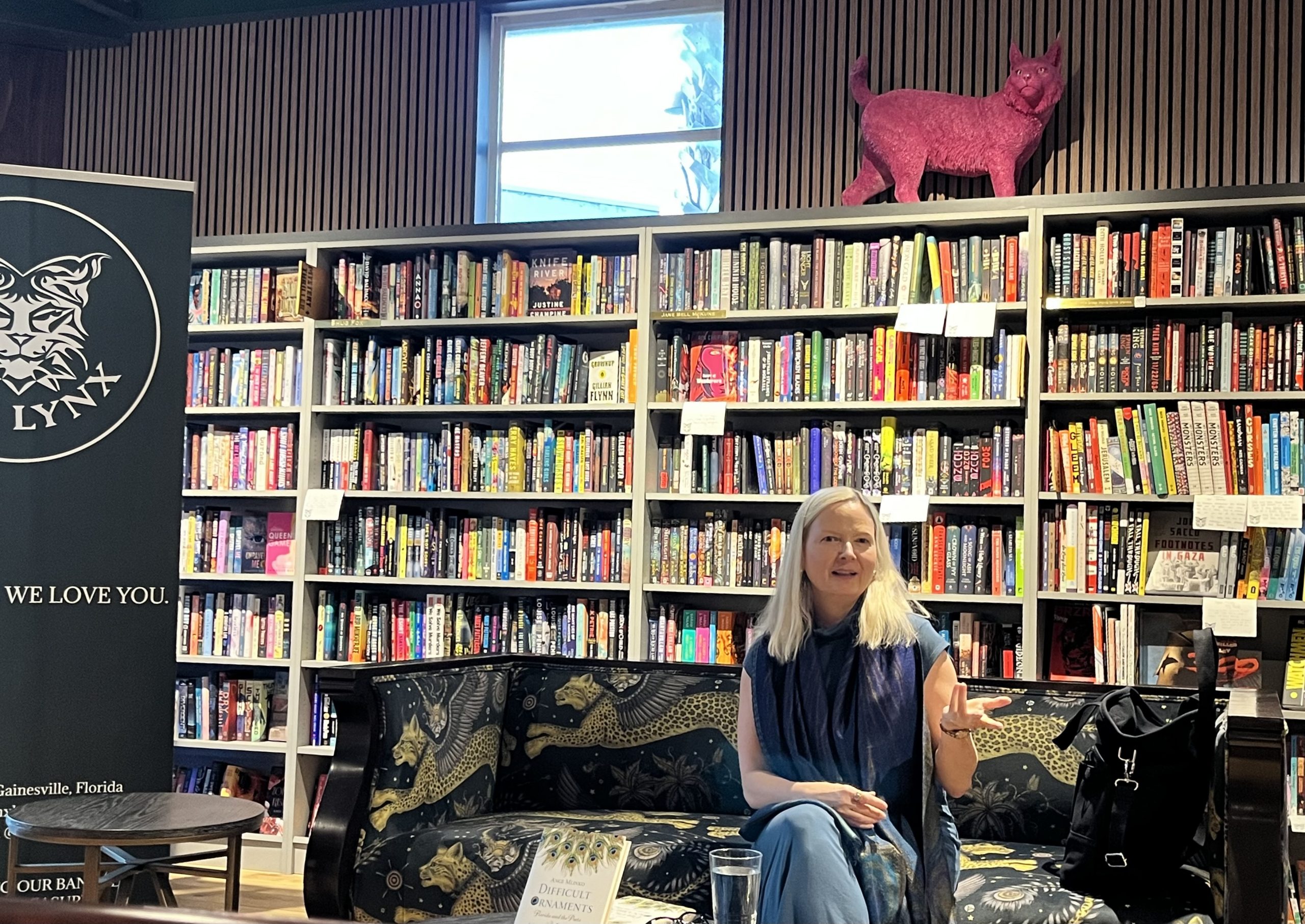 Ange Mlinko sits on a couch decorated in winged leopards. Behind her is walls of bookshelves. She is gesturing with her hand as though pictured mid-speech.