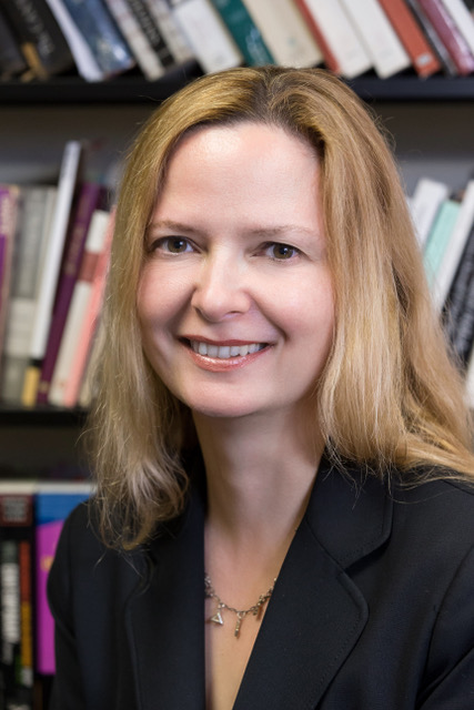 Professor Ange Mlinko smiles openly at the camera. She is wearing a black blazer and her long blonde hair rests on her shoulders. There is a full bookshelf visible in the background.
