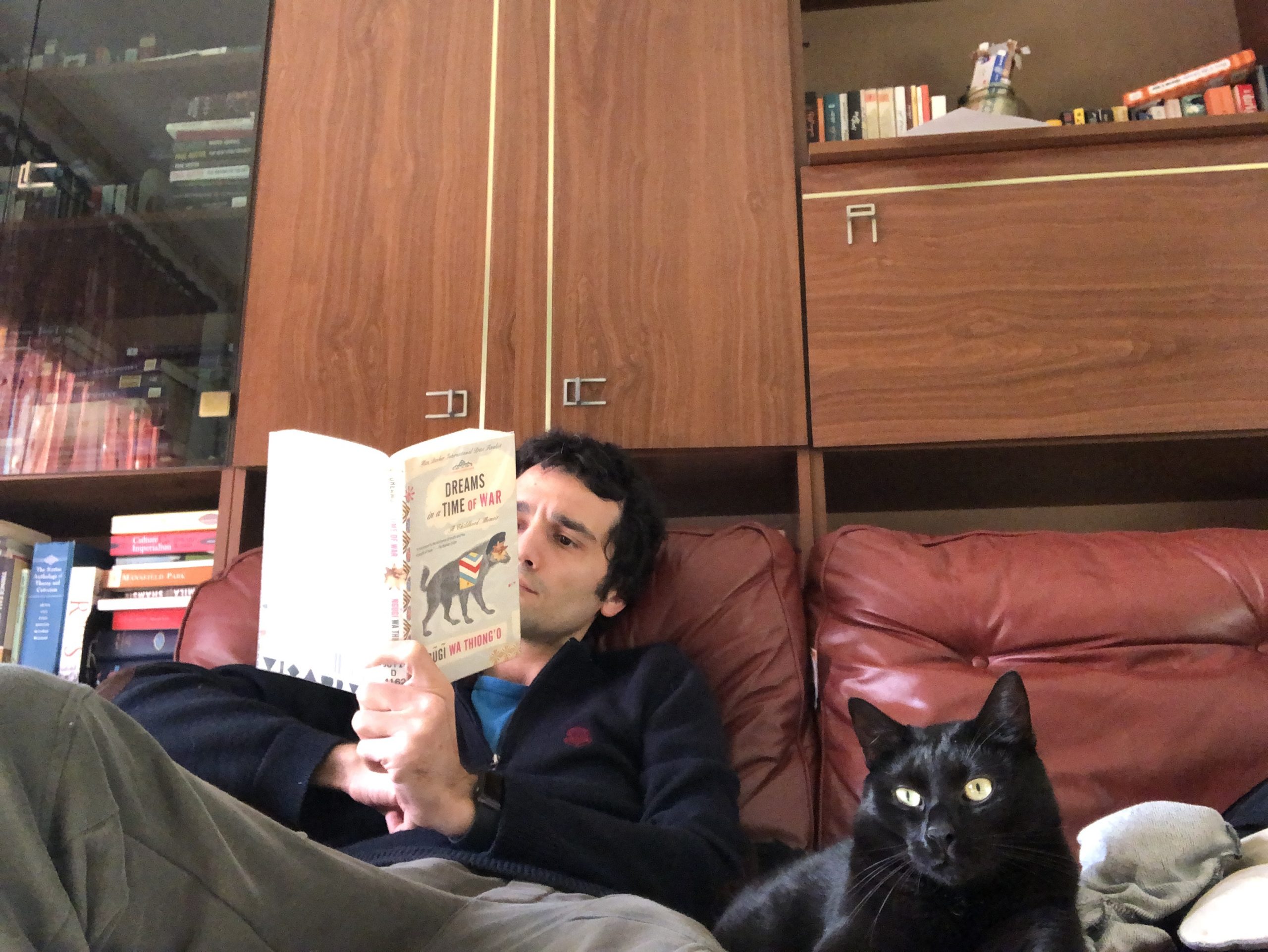 Armin Niknam is pictured seated on a red couch, reading beside his black cat, Fyodor.