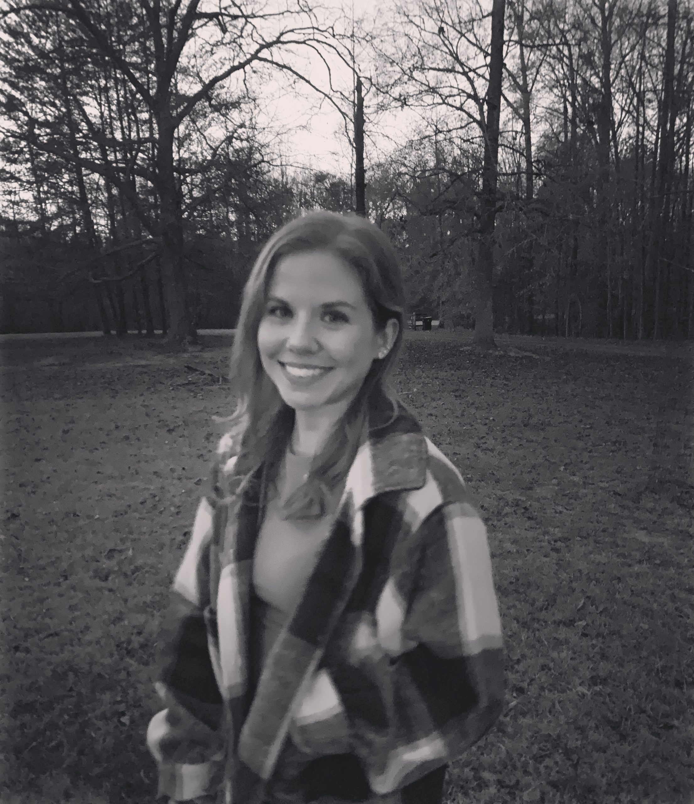 A black and white photo of Claire Beth Karnap standing in front of a picturesque landscape, with trees and spacious grass. She is wearing a plaid coat and smiles at the camera..