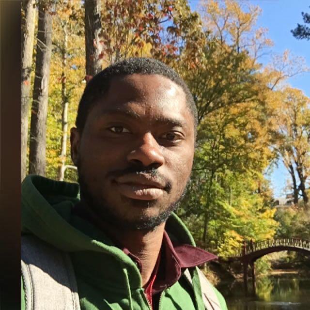 Cristovão Nwachukwu is pictured wearing a green jacket among picturesque autumnal scenery. Behind him is a small bridge over water and trees with yellow and red leaves. He is attending the 2019 Association for the Study of the Worldwide African Diaspora Conference