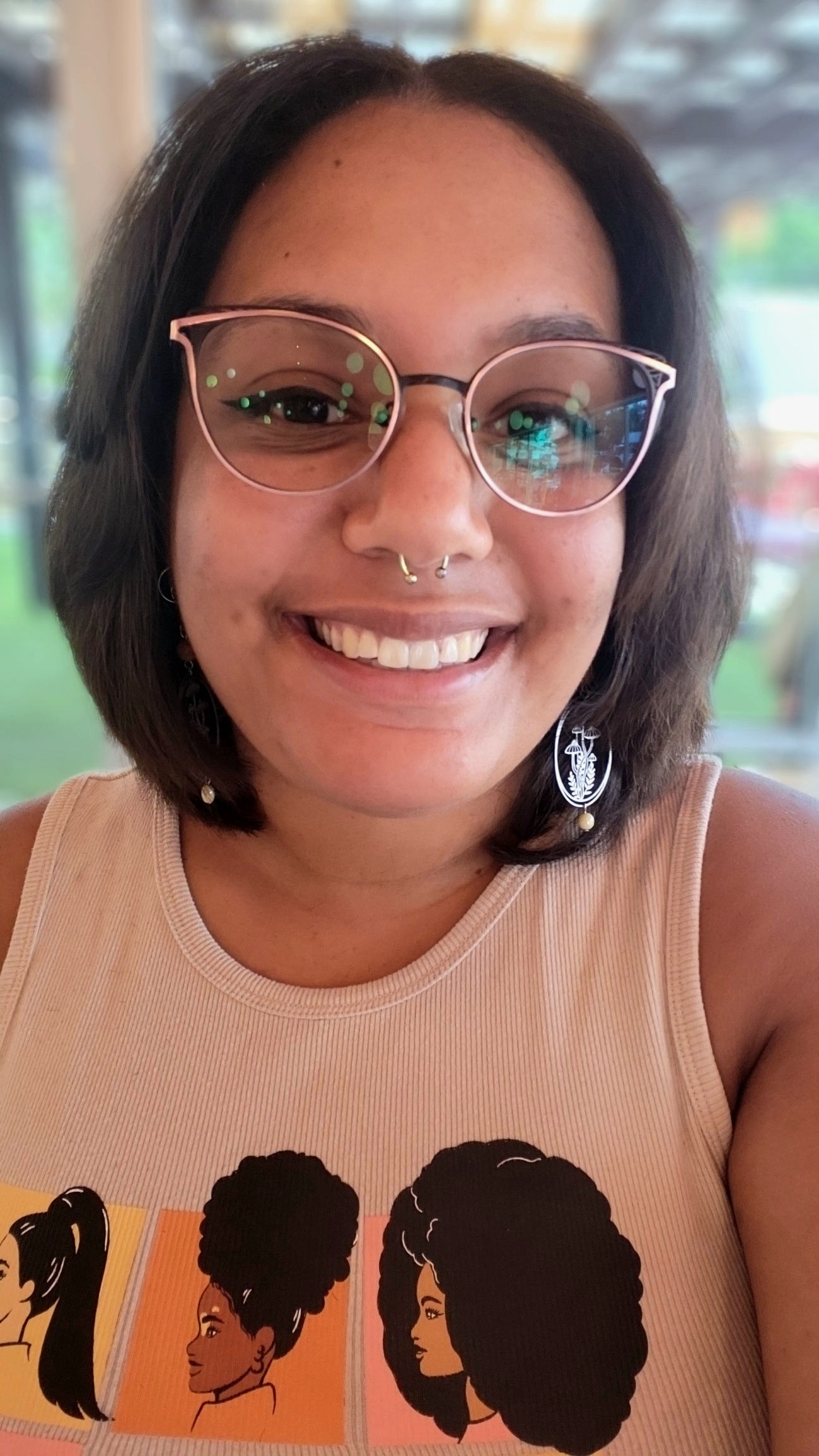 C.R. Dean smiles openly at the camera. She is wearing winged black eyeliner, glasses with black and rose gold metal rims, silver earrings that depict mushrooms and foliage, and a septum piercing. She also wears a light-colored tank top illustrated with the side profiles of Black women with different hairstyles.