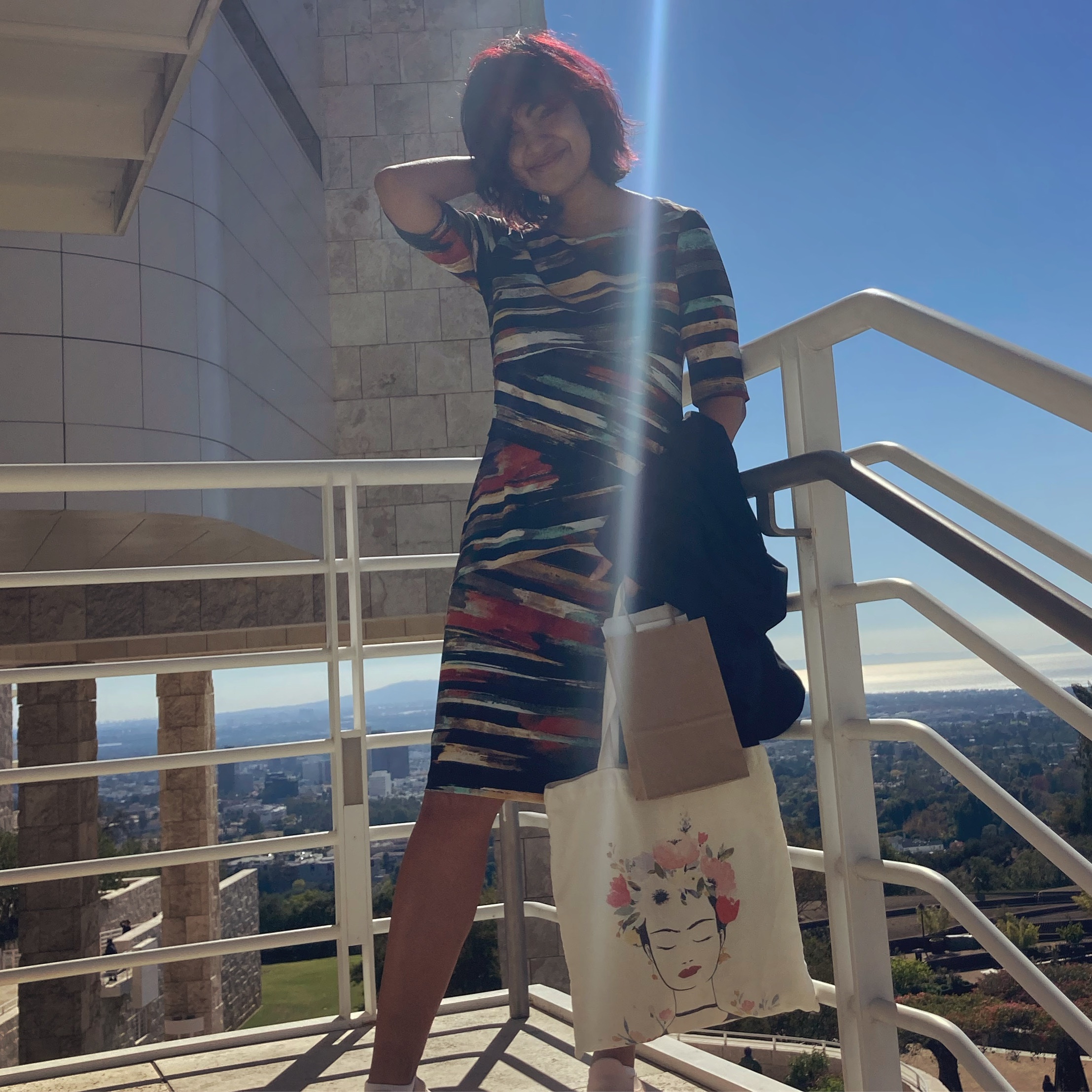 Debakanya is standing on a railed stairway by the water. She is wearing a black dress with colorful, abstractly applied stripes, and is carrying an illustrated tote bag. Her right hand is behind her head and she is smiling.