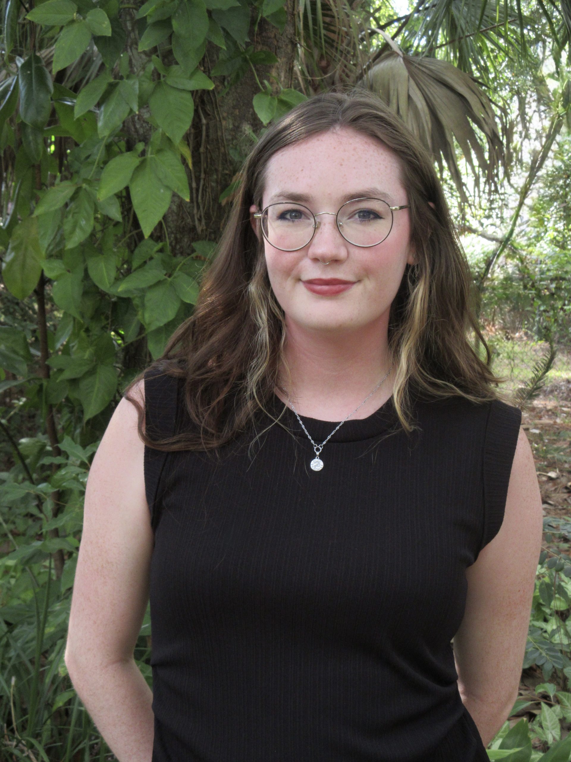 Ella Bissonnette smiles at the camera. She has brown hair, round metal glasses, and is wearing a black top. She is standing in front of a tree covered in green, leafy vines.