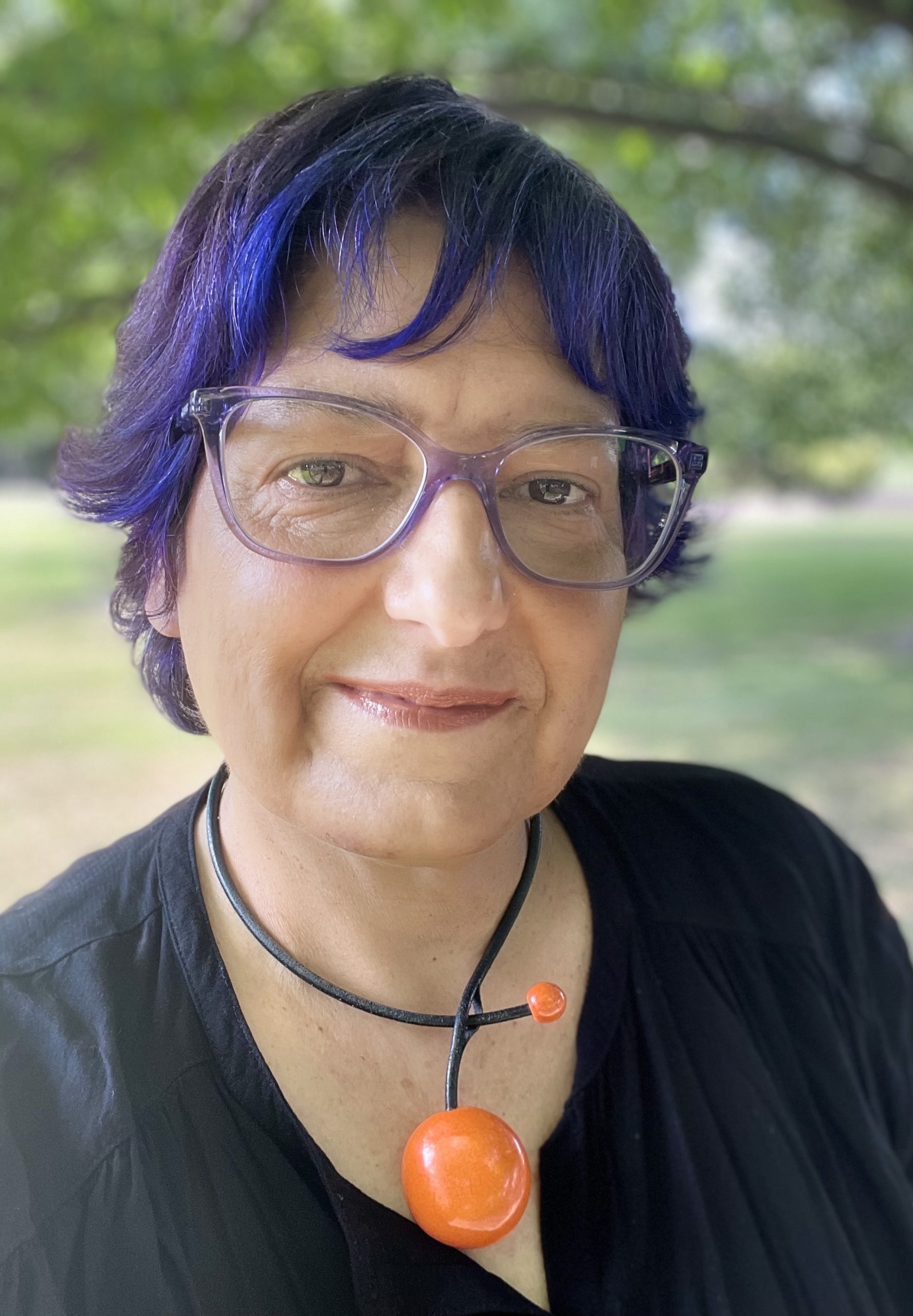 A headshot of Dr. Jodi Schorb, who is wearing glasses with translucent purple frames to match her hair, which is dyed purple. She is also wearing a black blouse and orange necklace, while smiling at the camera.