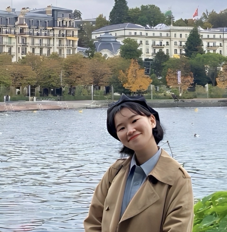 Hyunjung in Lozanne, France. She is wearing a light blue collared shirt under a trench coat and smiling. Behind her is a river, trees, and multistory buildings.