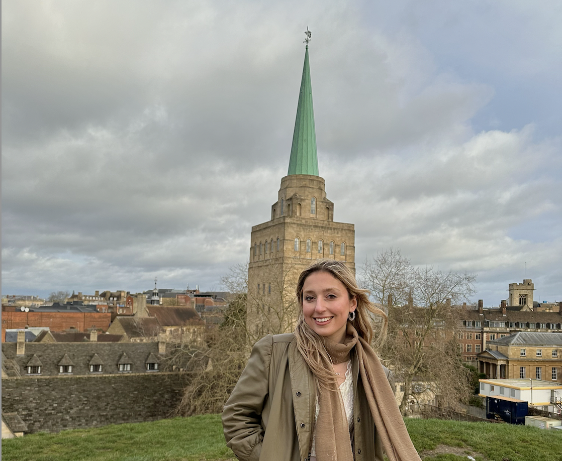 A photograph of Hilda standing on a rooftop terrace in front of a tower. She is smiling at the camera, wearing neutral tones and a scarf.