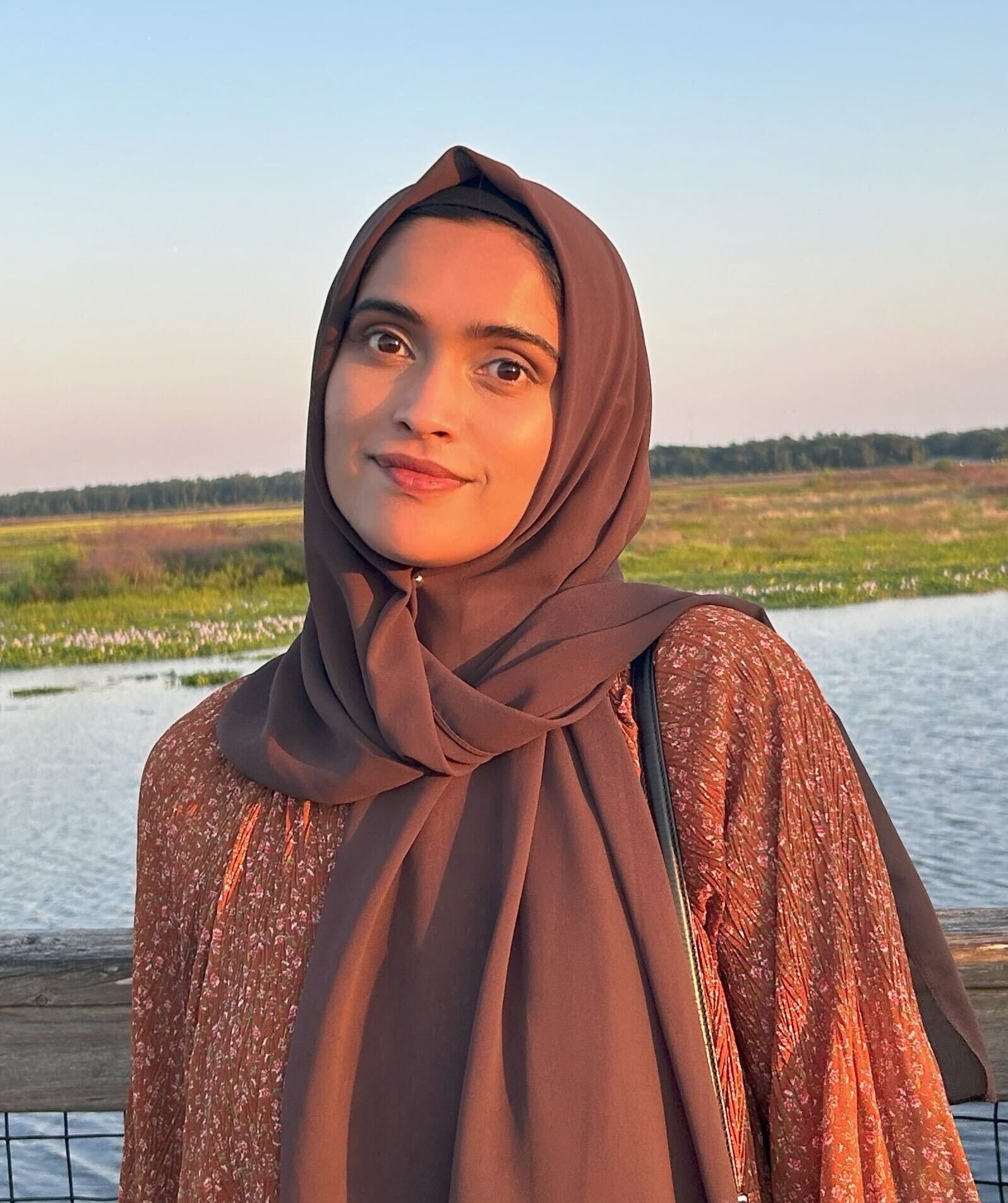 Iesha smiles softly at the camera. She is wearing a dark brown headscarf and orange top, standing in front of a lake.