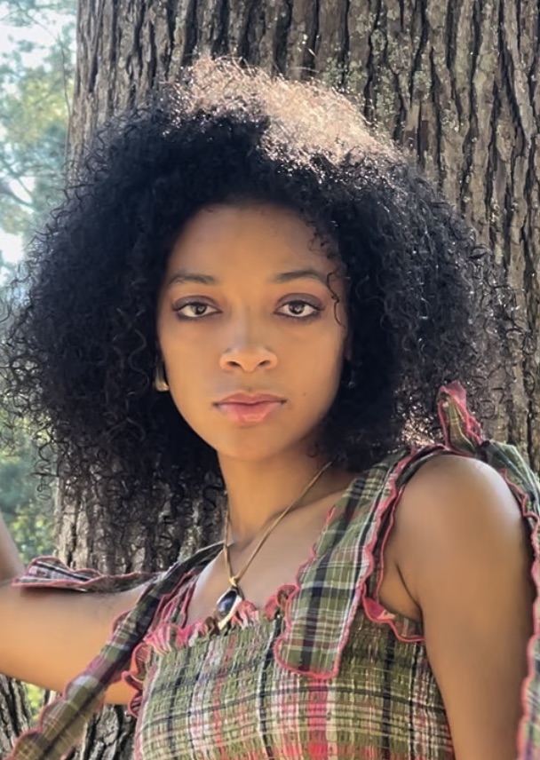 Israel stands in front of a tree and looks straight into the camera. She is wearing a green and pink plaid dress and a gold necklace. Her hair is dark, curly, and voluminous, stopping at her shoulders.