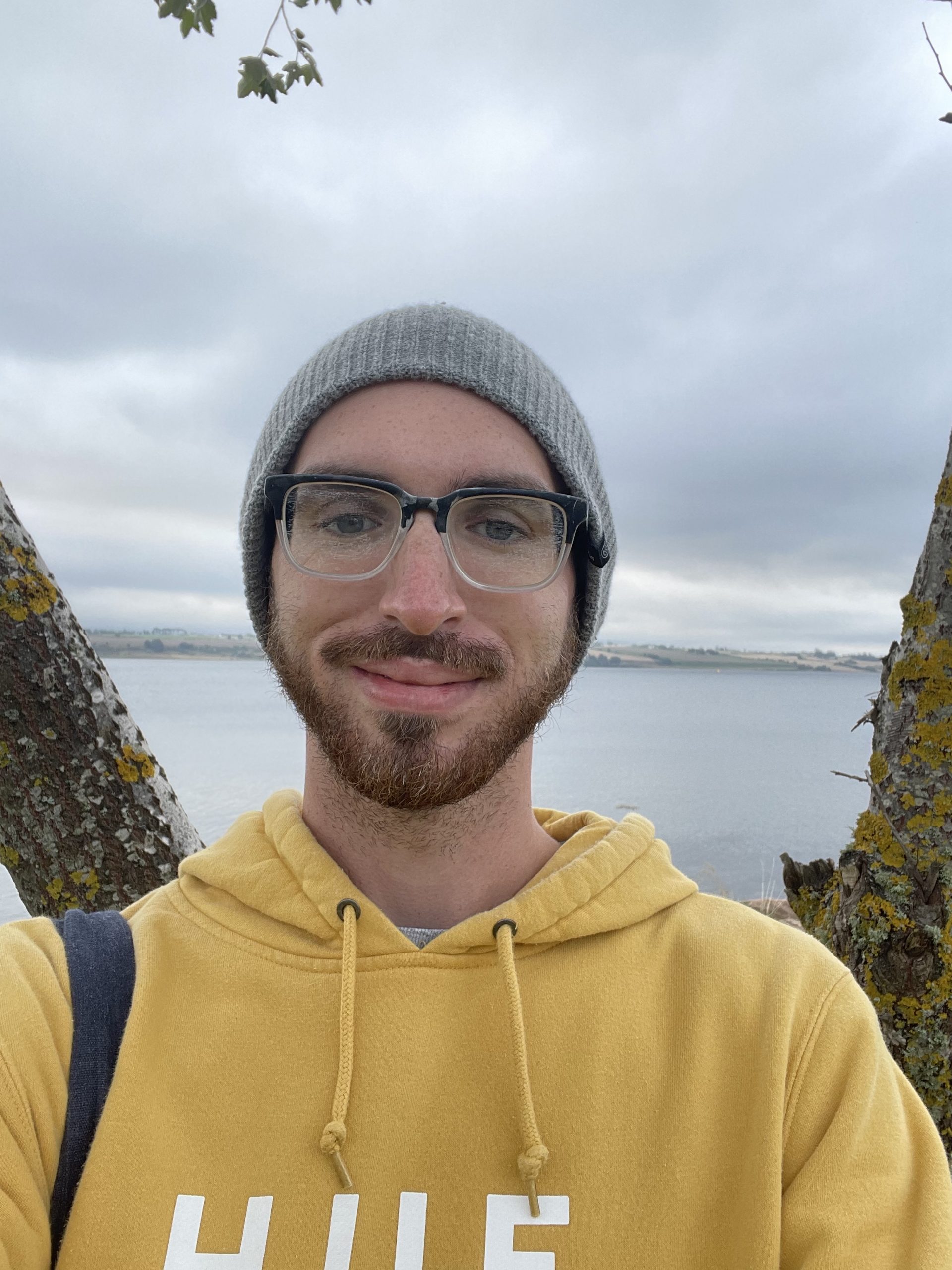 Jason Walker, wearing a yellow sweatshirt, grey beanie, and glasses, stands in front of open water.