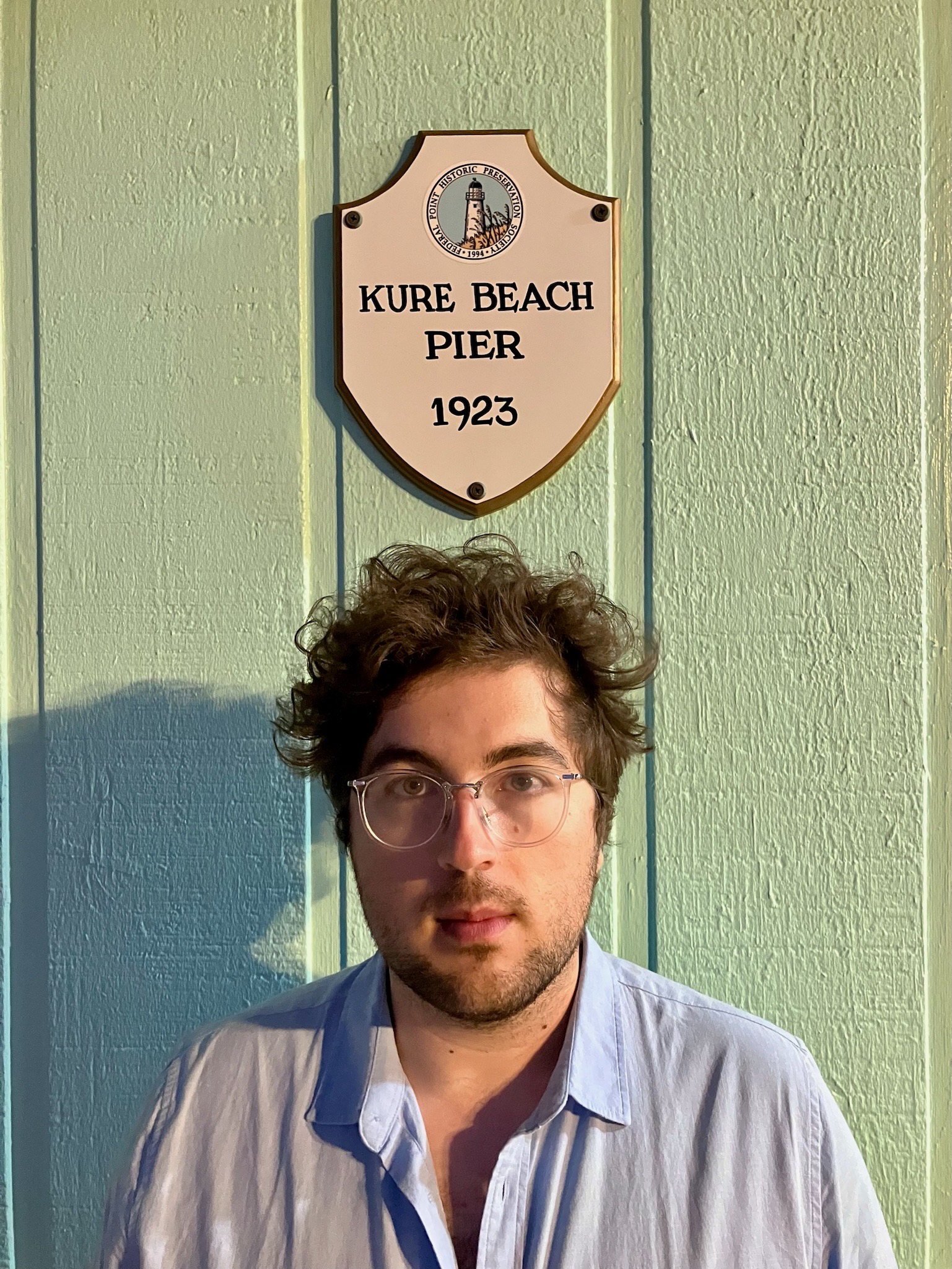 Jasey Roberts under a sign for the Kure Beach pier in North Carolina. They are wearing a blue collared shirt.