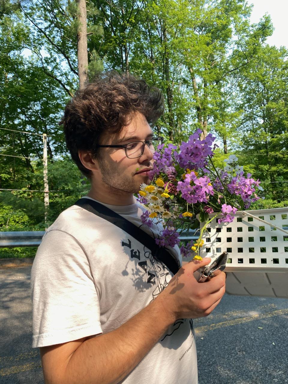 Jonathan, holding a basket of flowers with lowered eyes. He has brown hair and is wearing a white T-shirt