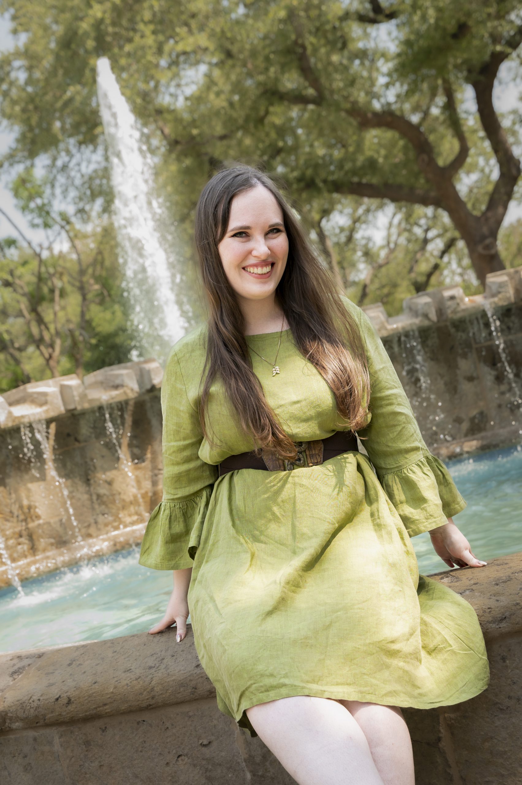 Kathryn smiles at camera while leaning against a fountain. She is wearing a green dress with a brown belt.