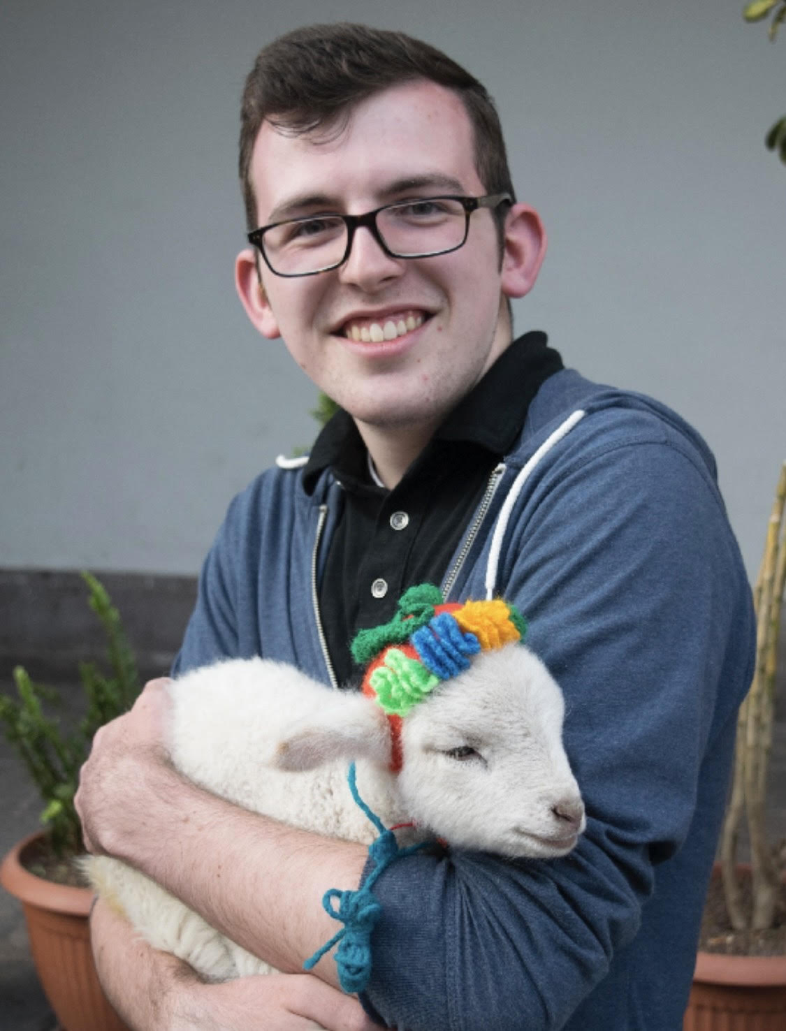William, smiling, holds a lamb on a street in Peru. The lamb is wearing a knitted hat.
