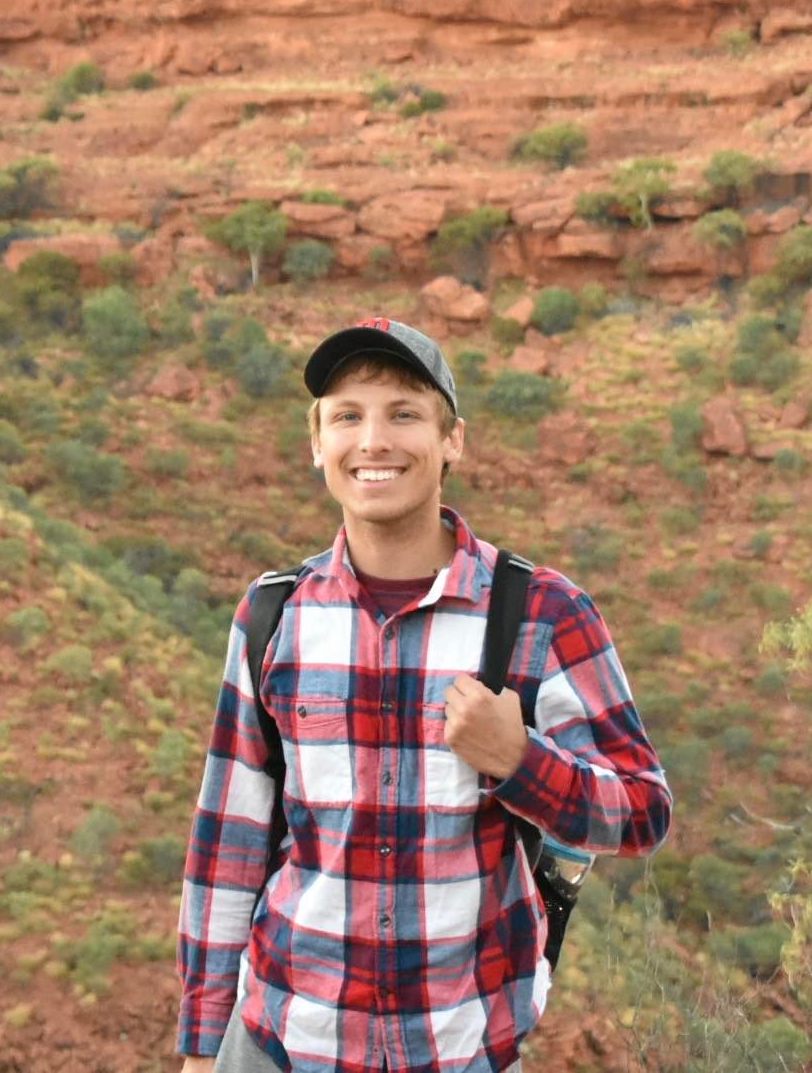 Luke smiles at the camera. They are wearing a red shirt with a baseball hat, in front of a canyon's rocky rim.