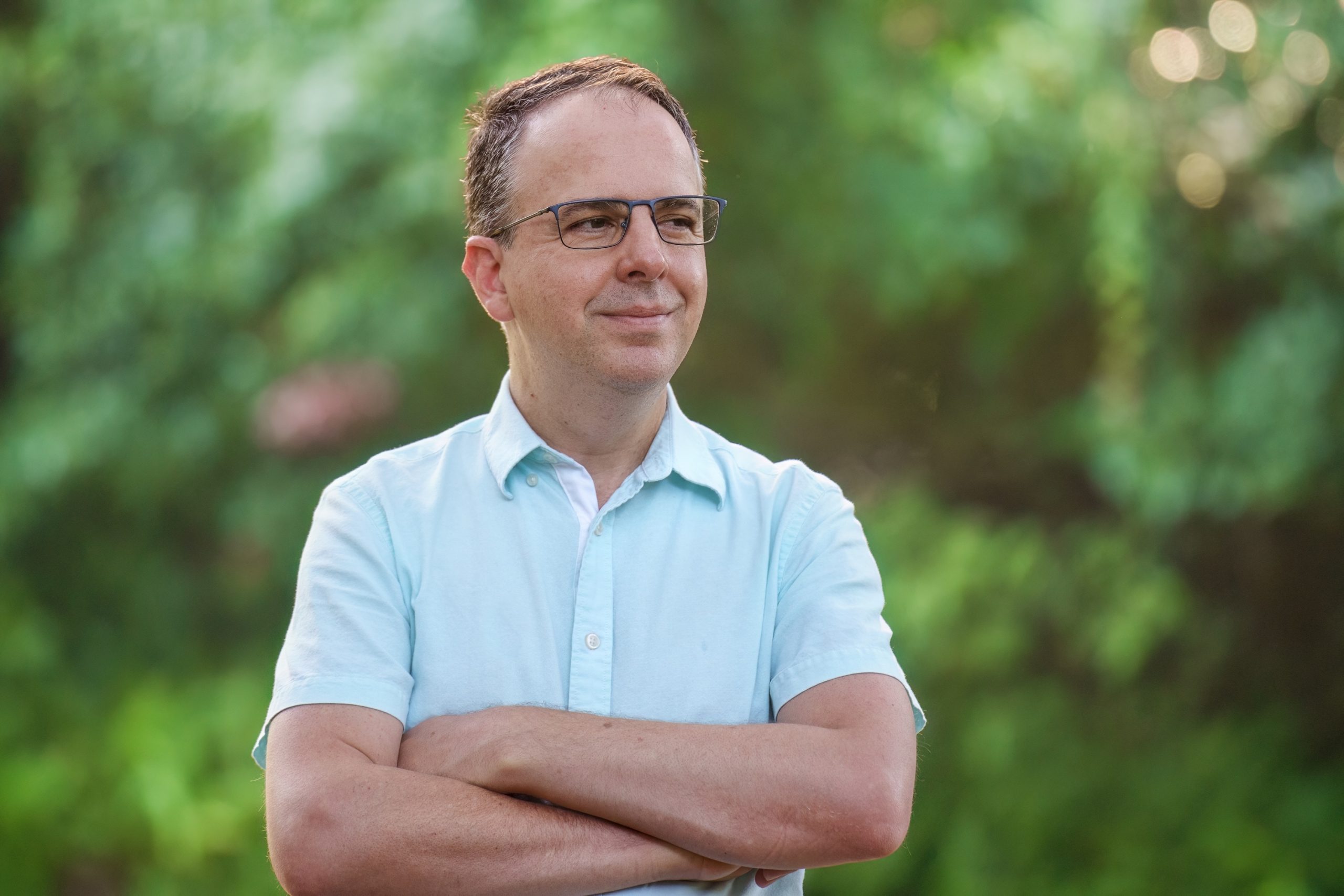 A headshot of Professor Maioli, who is looking off-camera and smiling with his arms crossed. He is wearing glasses and a light blue, button-up shirt with short sleeves. 